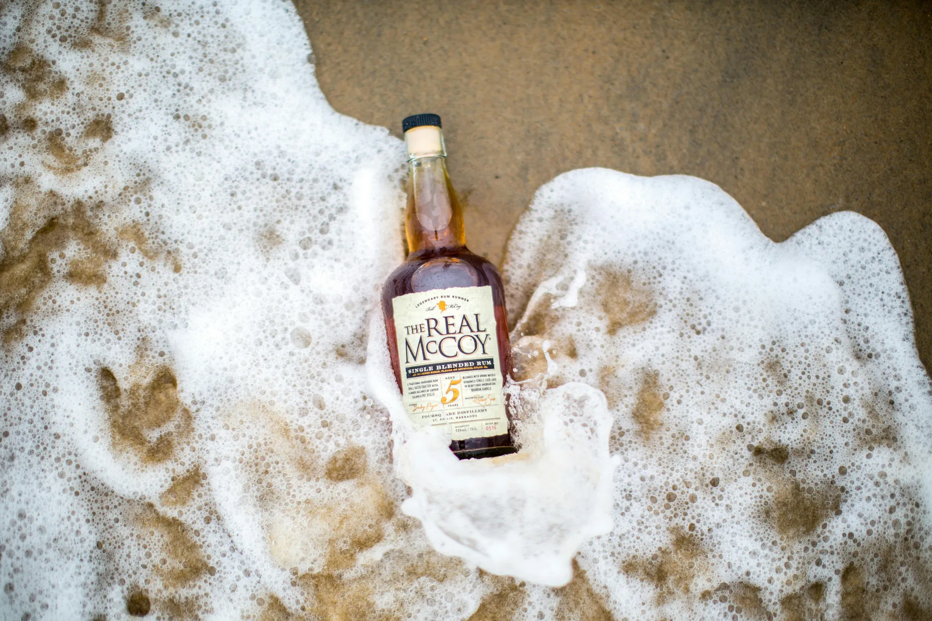 A bottle of The Real McCoy Rum lies in ocean foam on the beach.
